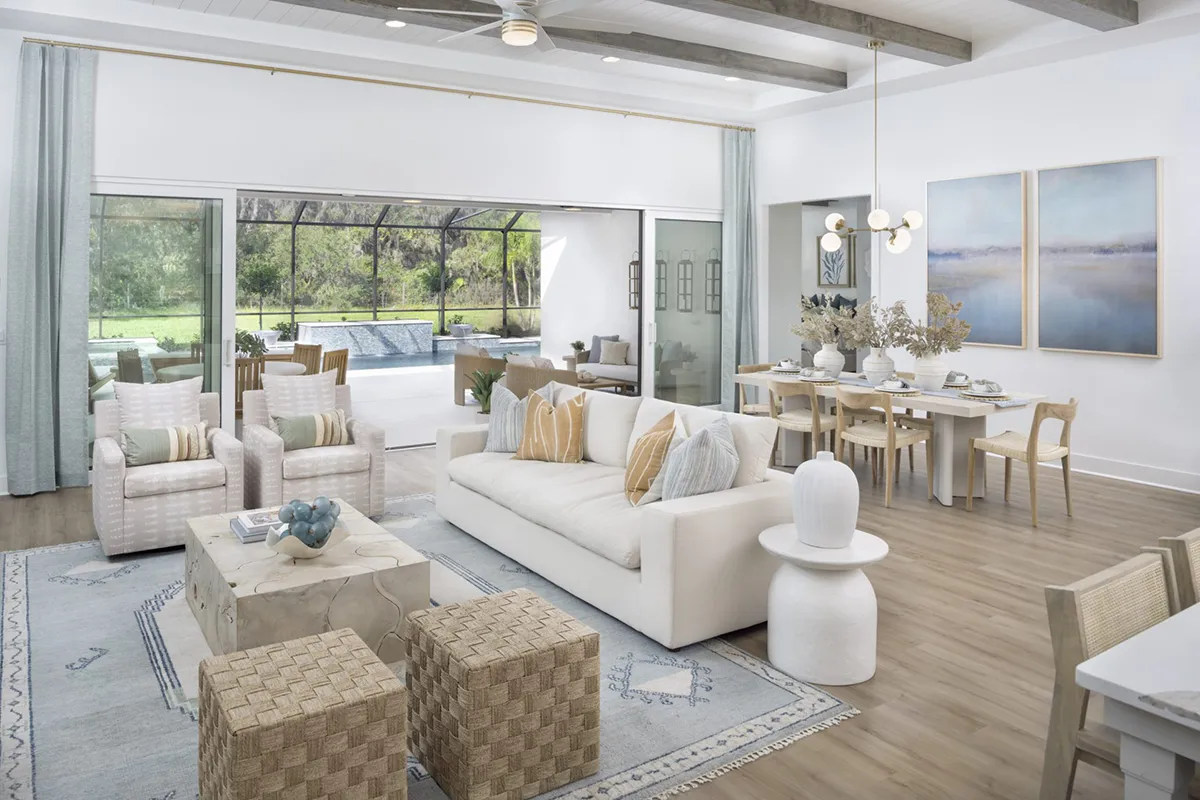 Modern open-concept living and dining area with light wood floors, neutral-toned furniture, large windows, and sliding glass doors opening to a patio with a pool and greenery outside. Decor is minimalist with natural accents.