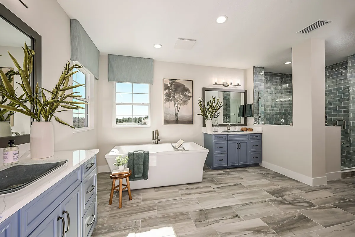 Modern bathroom with a freestanding tub, blue vanities, large mirrors, plants, and gray tile flooring. A window lets in natural light, and a glass-enclosed shower is visible in the background.