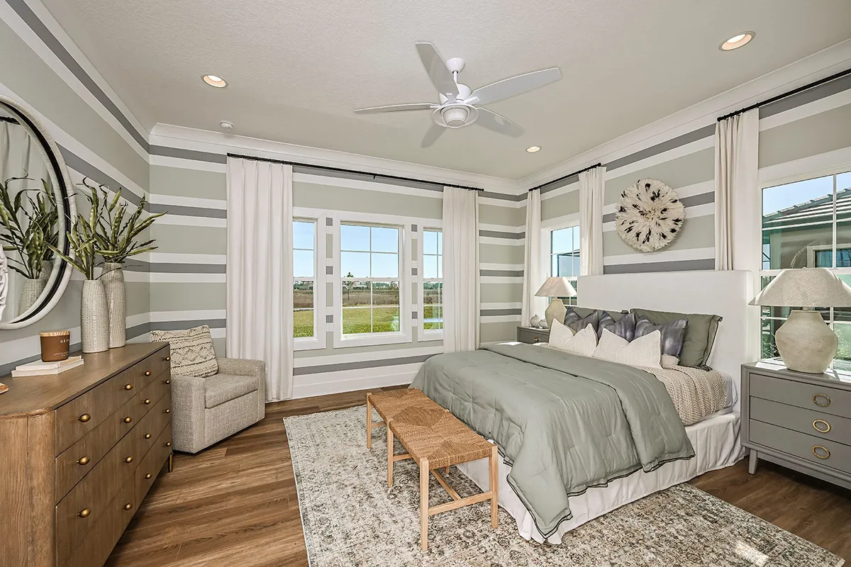 A modern bedroom with a queen bed, sage green bedding, and striped walls. There are large windows with white curtains, a wooden dresser with a round mirror, two nightstands with lamps, and a woven bench at the foot of the bed.