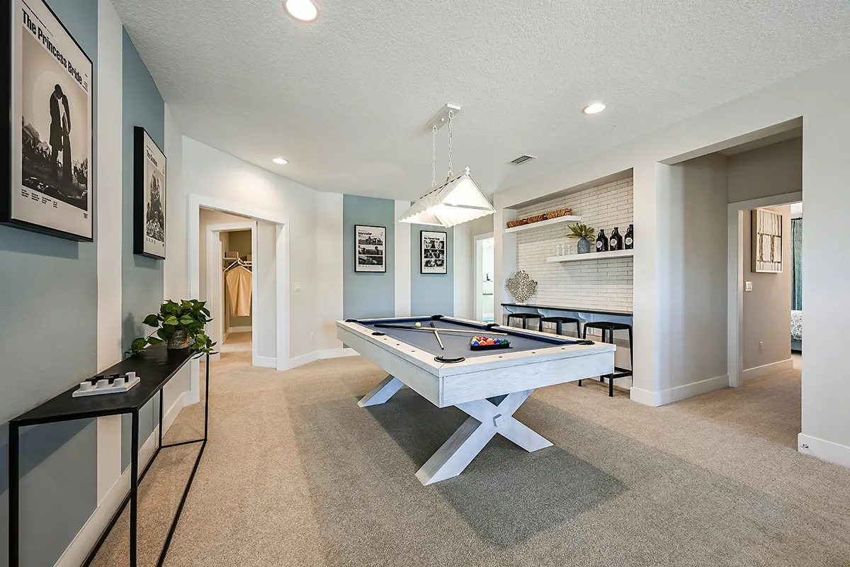 A modern game room with a white pool table in the center, cues on the wall, framed artwork, a small bar with shelves, neutral walls, beige carpet, and contemporary decor.