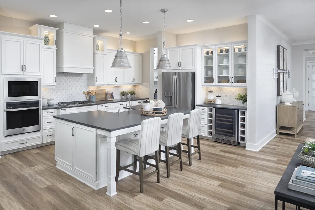 Modern kitchen with white cabinets, stainless steel appliances, a large island with three upholstered bar stools, pendant lights, and wood-look flooring. Glass-front cabinets and decorative items add a stylish touch.
