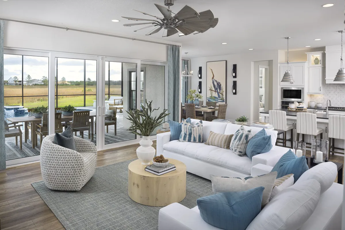 A modern living room with white sofas, blue accents, a round wooden coffee table, and large glass doors leading to an outdoor patio. An open kitchen and dining area are visible in the background.