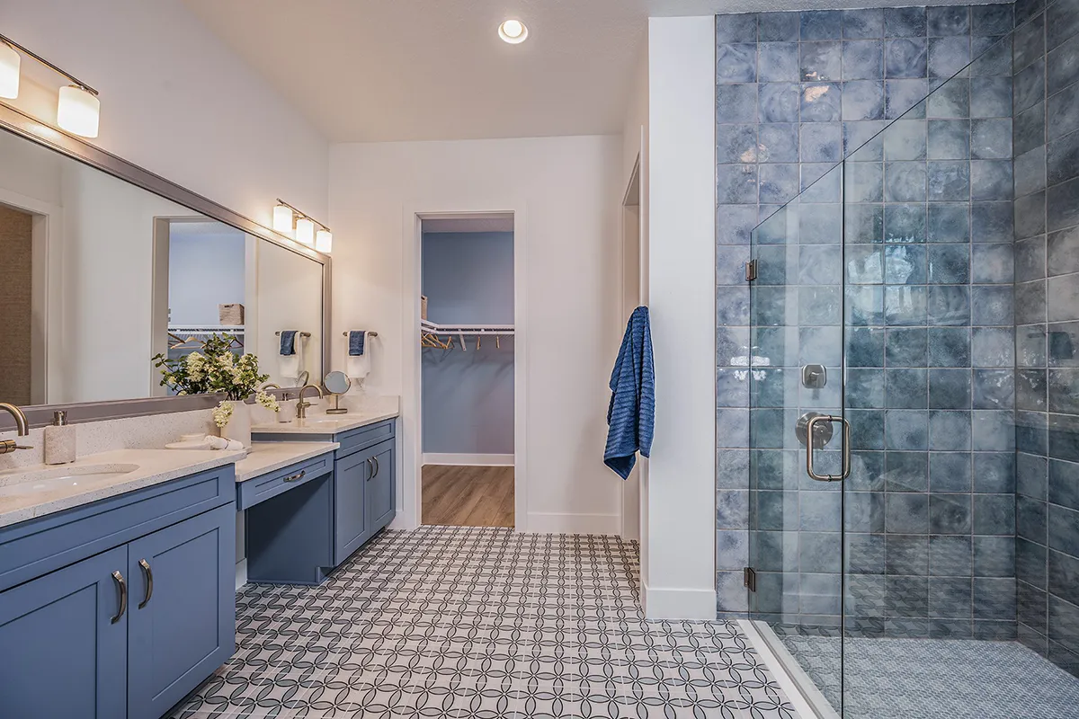 A modern bathroom with blue cabinets, double sinks, a large mirror, patterned floor tiles, and a glass walk-in shower with blue tiles. A closet with wooden flooring is visible through an open doorway.