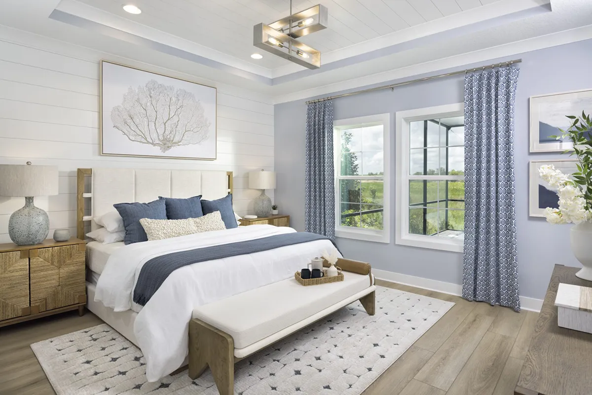 Bright, modern bedroom with a large bed featuring white and blue bedding, wooden nightstands with lamps, a bench at the foot of the bed, patterned curtains, and a large window letting in natural light.