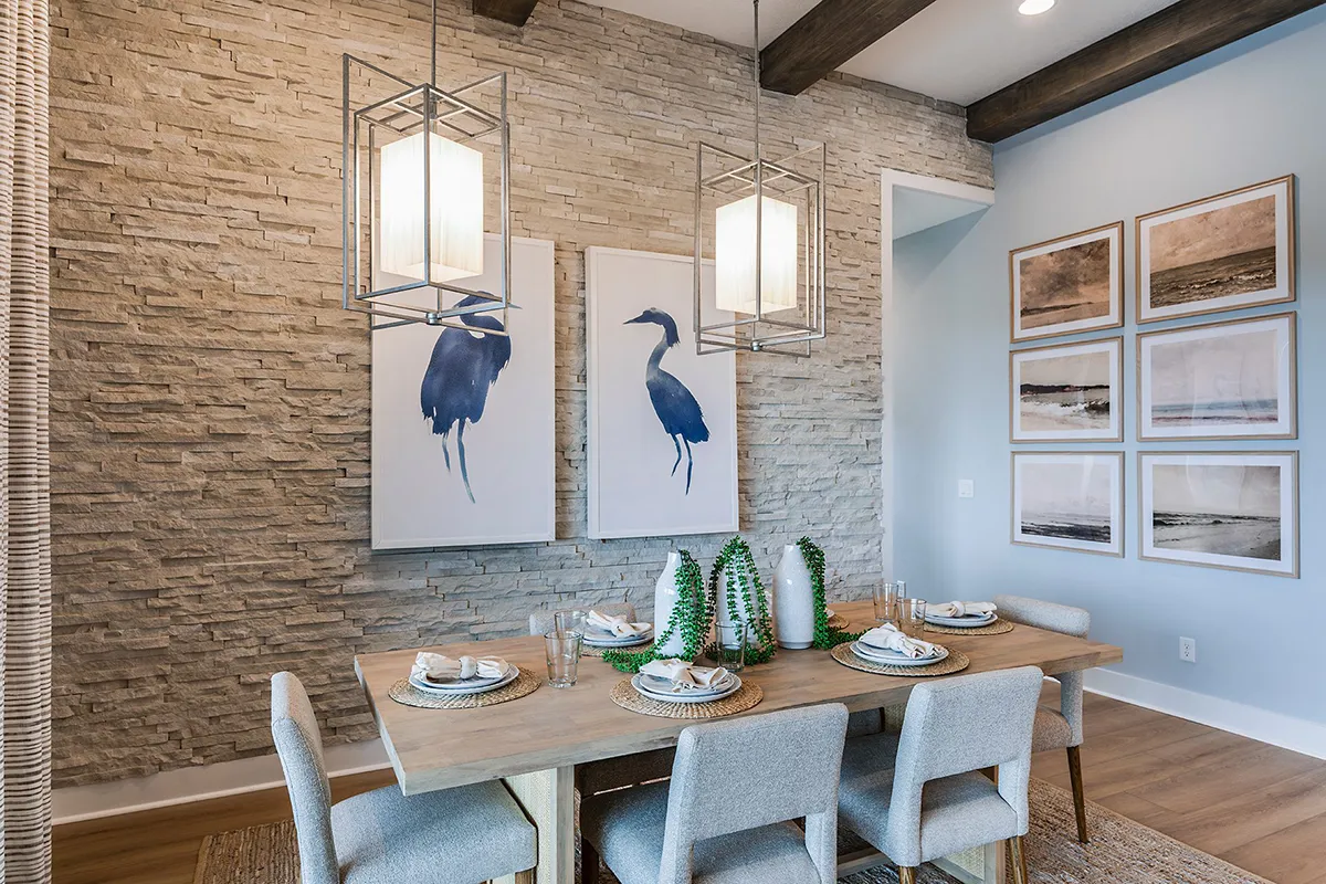 A modern dining room with a wooden table set for six, gray upholstered chairs, stone accent wall, two large bird paintings, geometric pendant lights, and framed art on a pale blue wall.