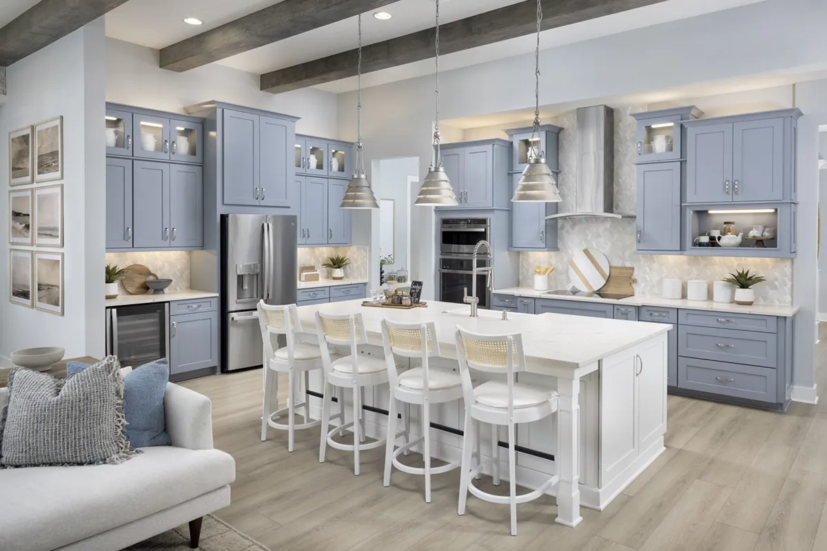 Modern kitchen with light blue cabinets, a large white island with four white chairs, stainless steel appliances, pendant lights, and light wood flooring. Decorative items and plants are arranged on the counters and shelves.