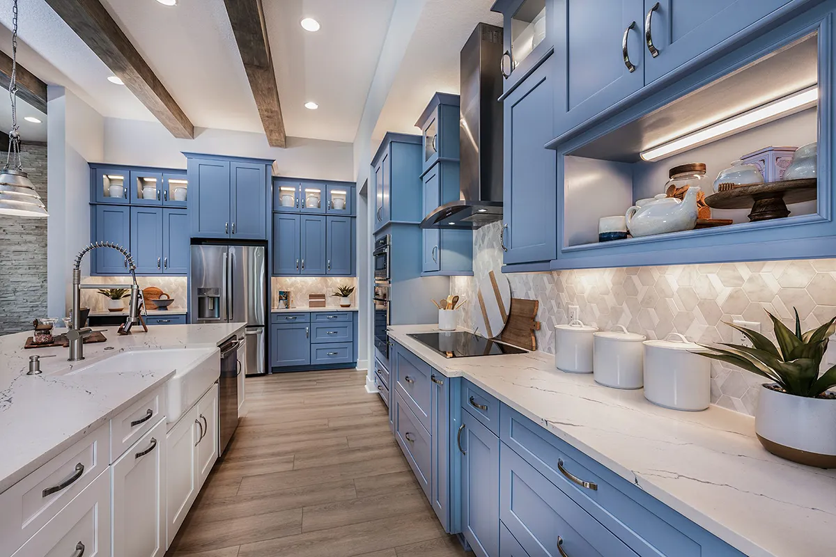 Modern kitchen with blue cabinets, white countertops, stainless steel appliances, a large island with a sink, wooden ceiling beams, under-cabinet lighting, and decorative dishes and plants on display.
