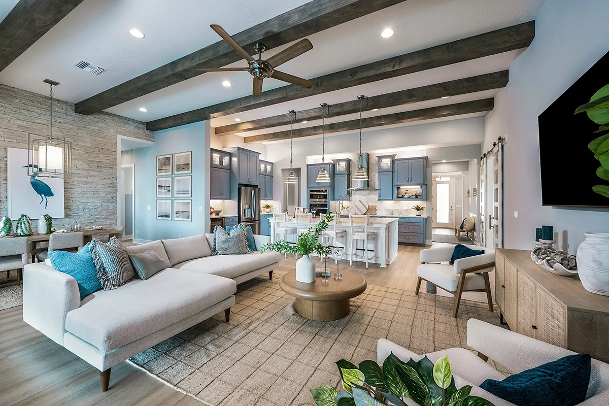 Modern open-concept living room and kitchen with exposed wood beams, light furniture, blue accents, pendant lighting, and large windows. Cosy seating area, decorative plants, and a spacious, welcoming atmosphere.