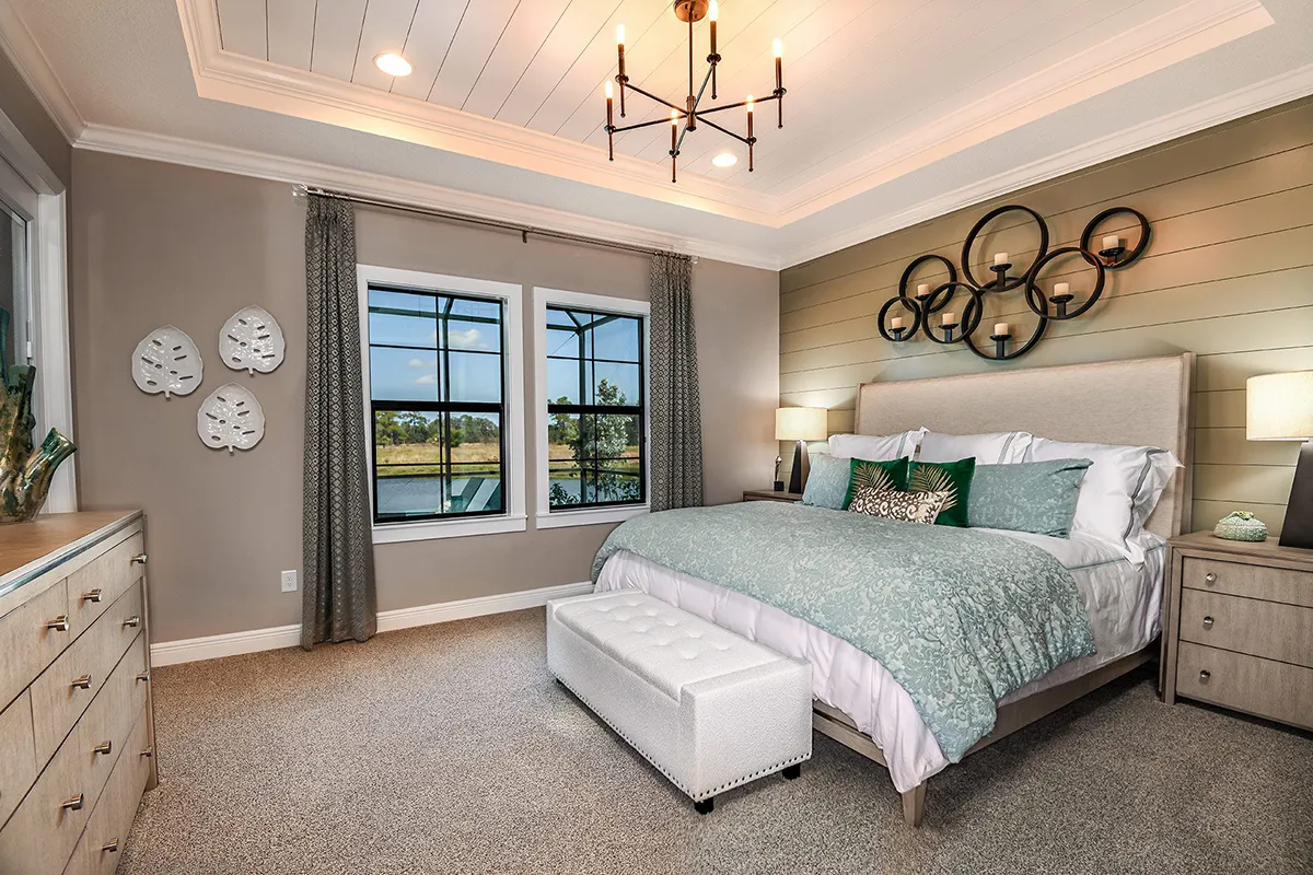 A modern bedroom with a large bed, green and white bedding, bedside lamps, a padded bench, wall art, a dresser, carpeted floor, and two large windows with views of greenery and water.