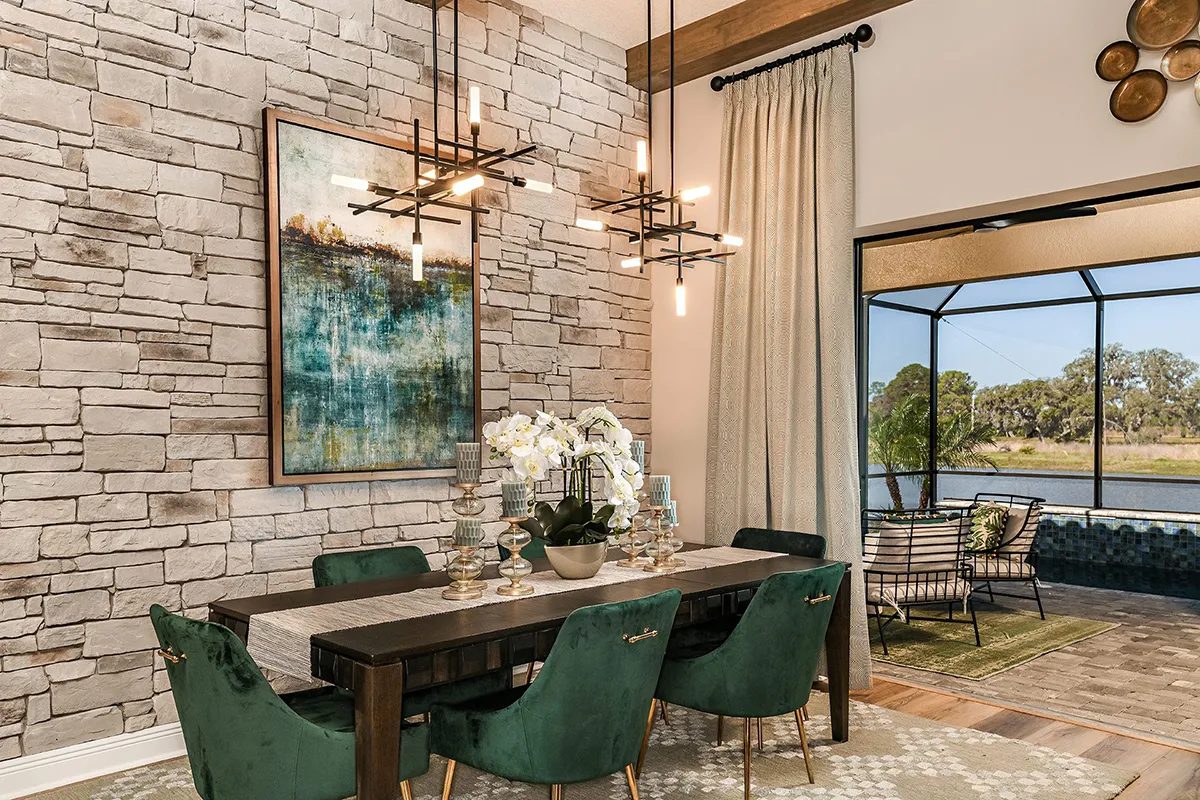 A modern dining room with a stone accent wall, abstract artwork, green velvet chairs, a wooden table with flowers, and a contemporary chandelier. Sliding glass doors open to a patio with outdoor seating and a view of trees.