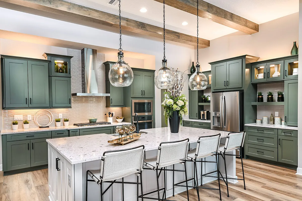 Modern kitchen with green cabinets, stainless steel appliances, a large white island with four barstools, pendant lights, wood ceiling beams, and decorative plants and vases.