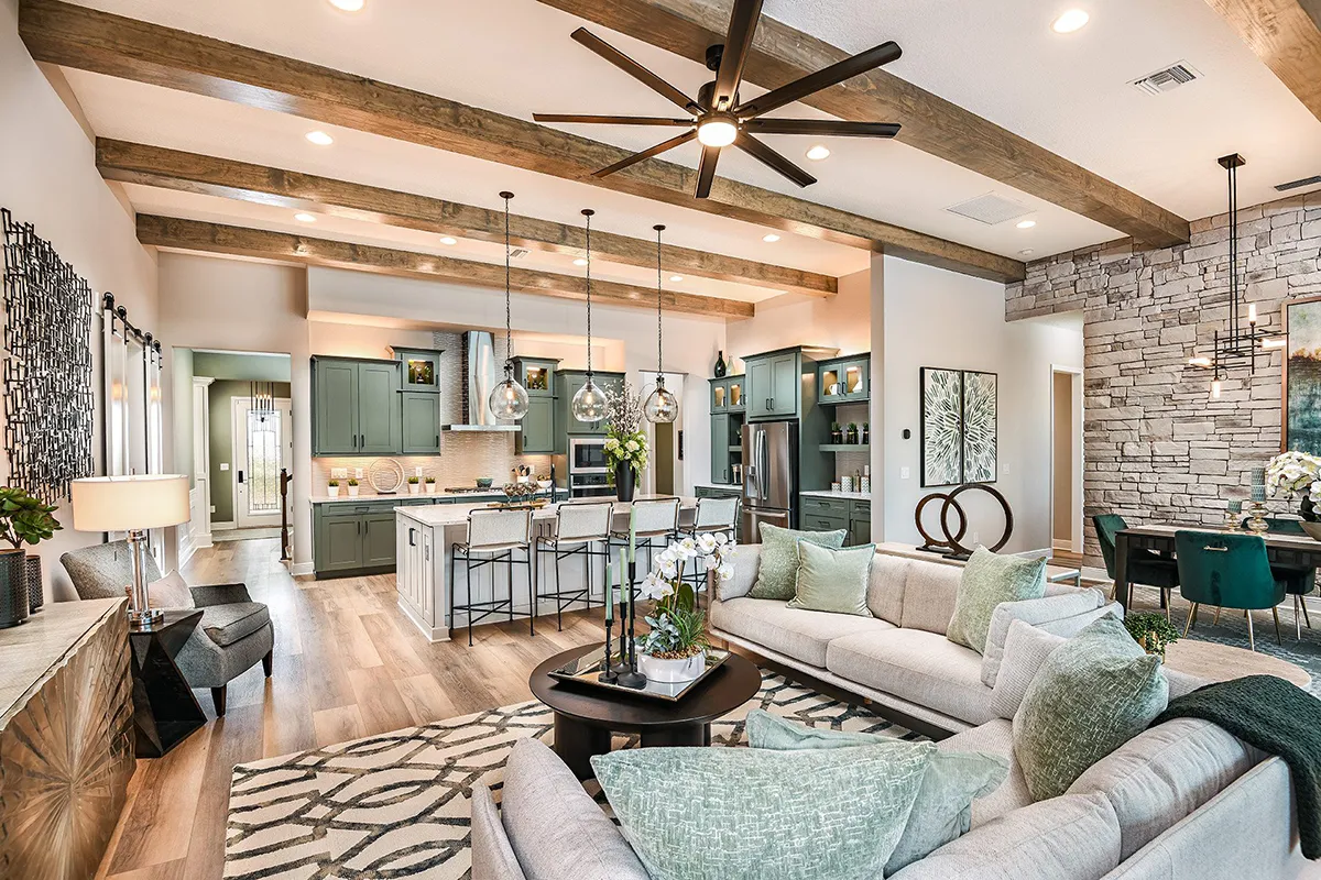 Spacious open-concept living area with exposed wood beams, modern kitchen with green cabinets, island and pendant lights, cozy sofas, round coffee table, dining area, and accent stone wall.
