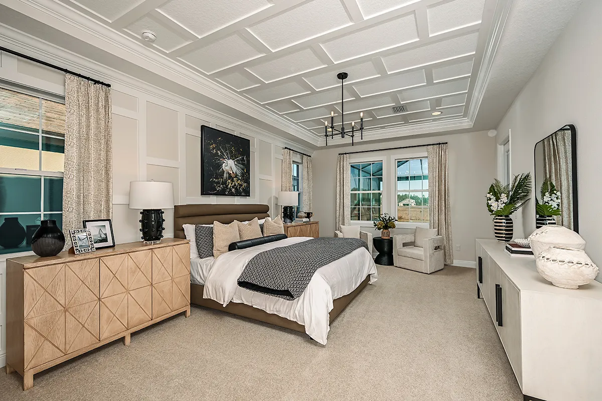 A modern, spacious bedroom with a large bed, patterned bedding, two wooden nightstands, lamps, a chandelier, armchairs by tall windows, light carpet, and neutral decor accents.