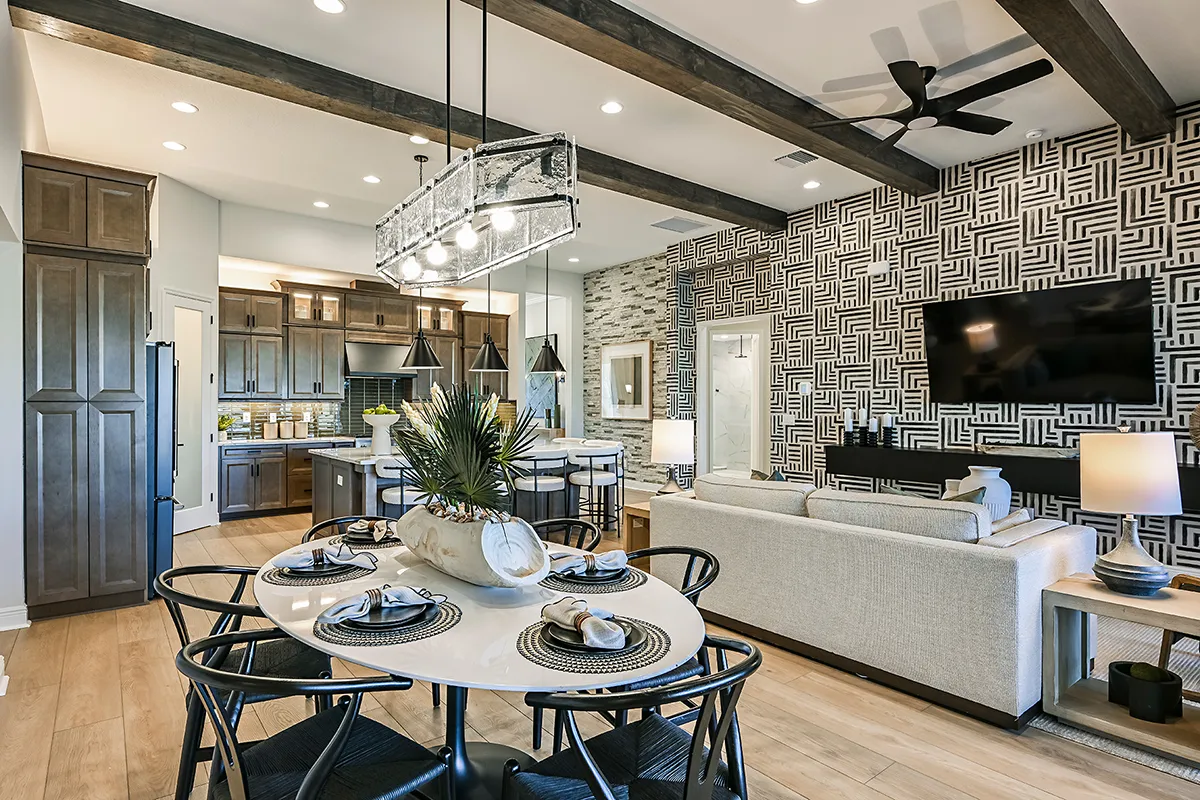 Modern open-concept kitchen and living room with wood accents, geometric wallpaper, a round dining table set for six, and a large wall-mounted TV above a light sofa. Exposed beams and pendant lighting complete the space.