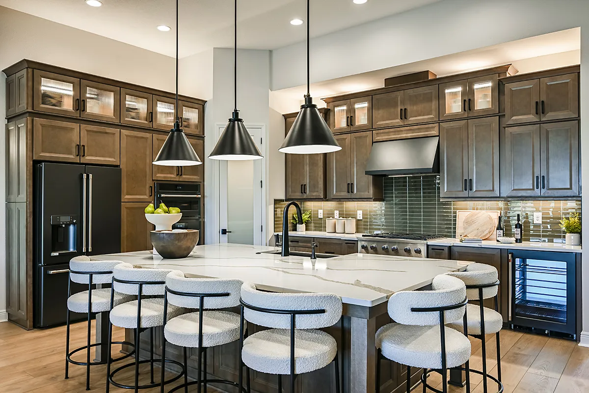 Modern kitchen with wooden cabinets, black appliances, a large white island with six cushioned barstools, three black pendant lights, a built-in oven, and glass door accents on upper cabinets.