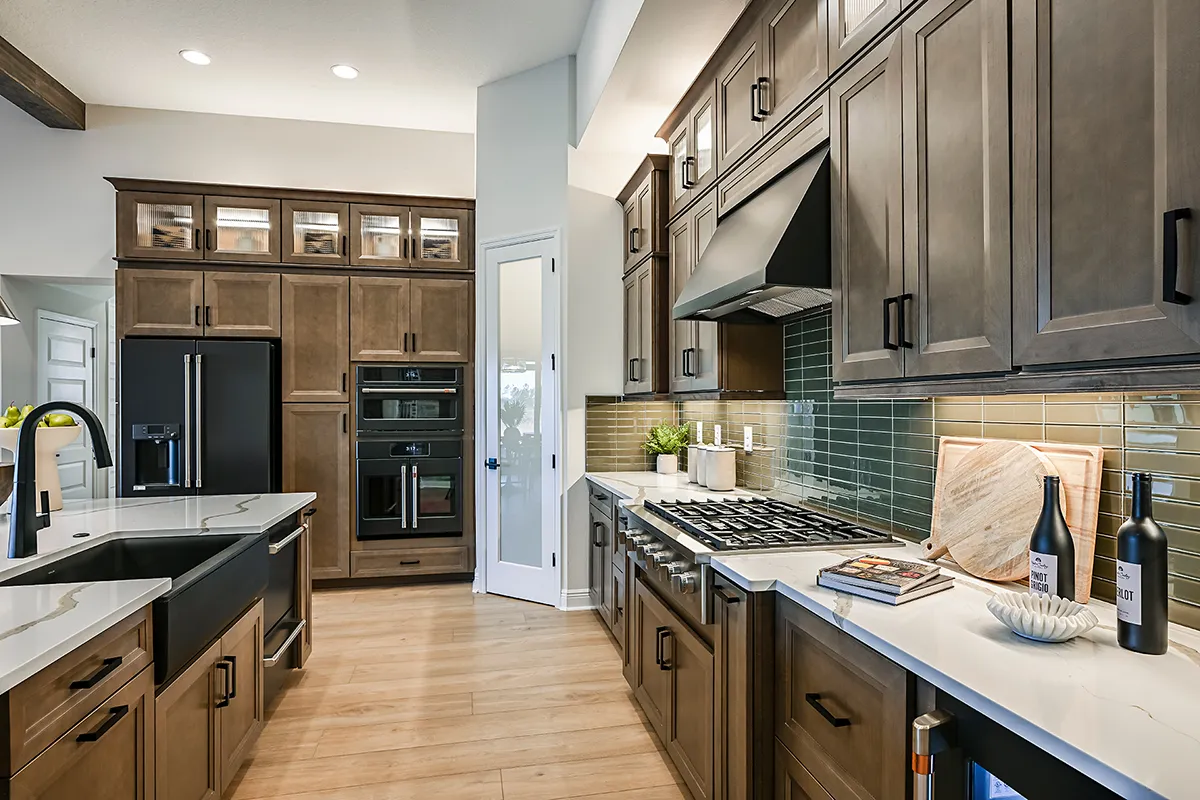 Modern kitchen with dark wood cabinets, stainless steel appliances, white countertops, a gas stove, black sink, and green glass tile backsplash. Items like wine bottles and cookbooks are on the counter.