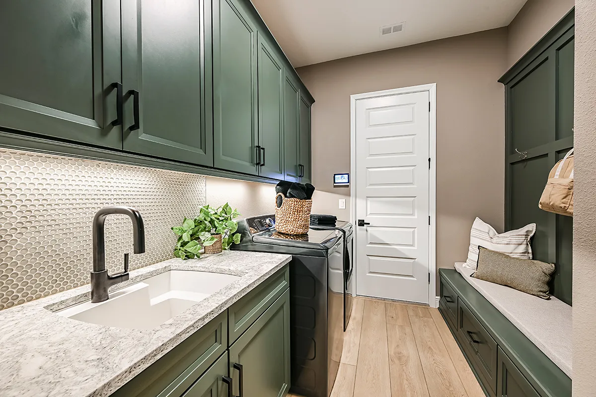 A modern laundry room with green cabinets, a white countertop and sink, a washing machine, a dryer, a bench with pillows, and a plant; light wood flooring and a white door are visible.