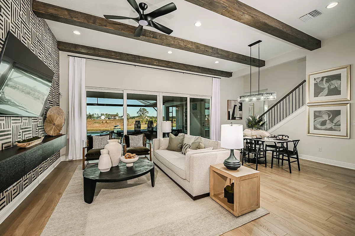 Modern living and dining area with wood beams, patterned accent wall, neutral furnishings, large glass doors leading to patio, and natural light streaming in. Black ceiling fan and contemporary decor complete the space.