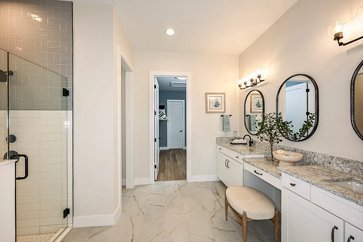 Modern bathroom with a glass shower, marble floor, white cabinetry, granite countertops, double sinks with oval mirrors, wall sconces, and a cushioned bench under the vanity. A hallway is visible through the doorway.
