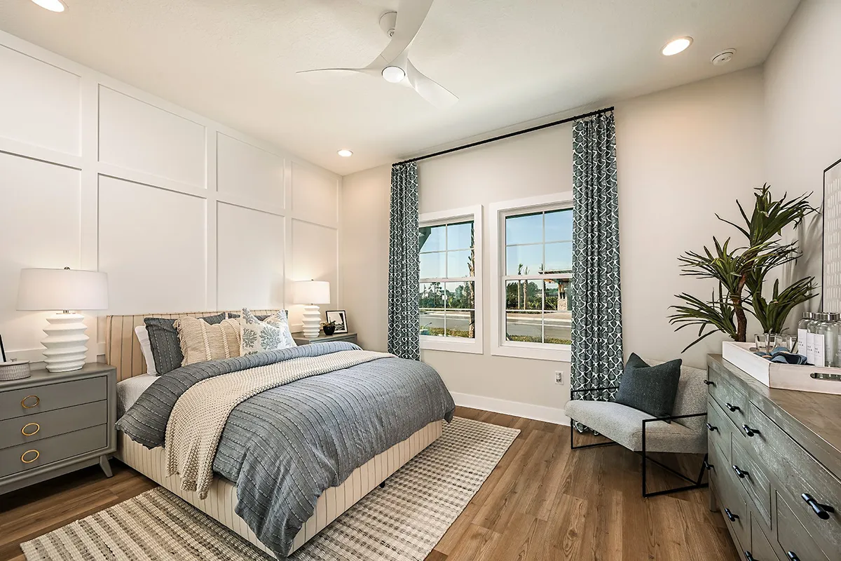 Modern bedroom with a large bed, gray and white bedding, two nightstands with lamps, a dresser, a gray chair, houseplants, wood flooring, and two windows with patterned curtains letting in natural light.