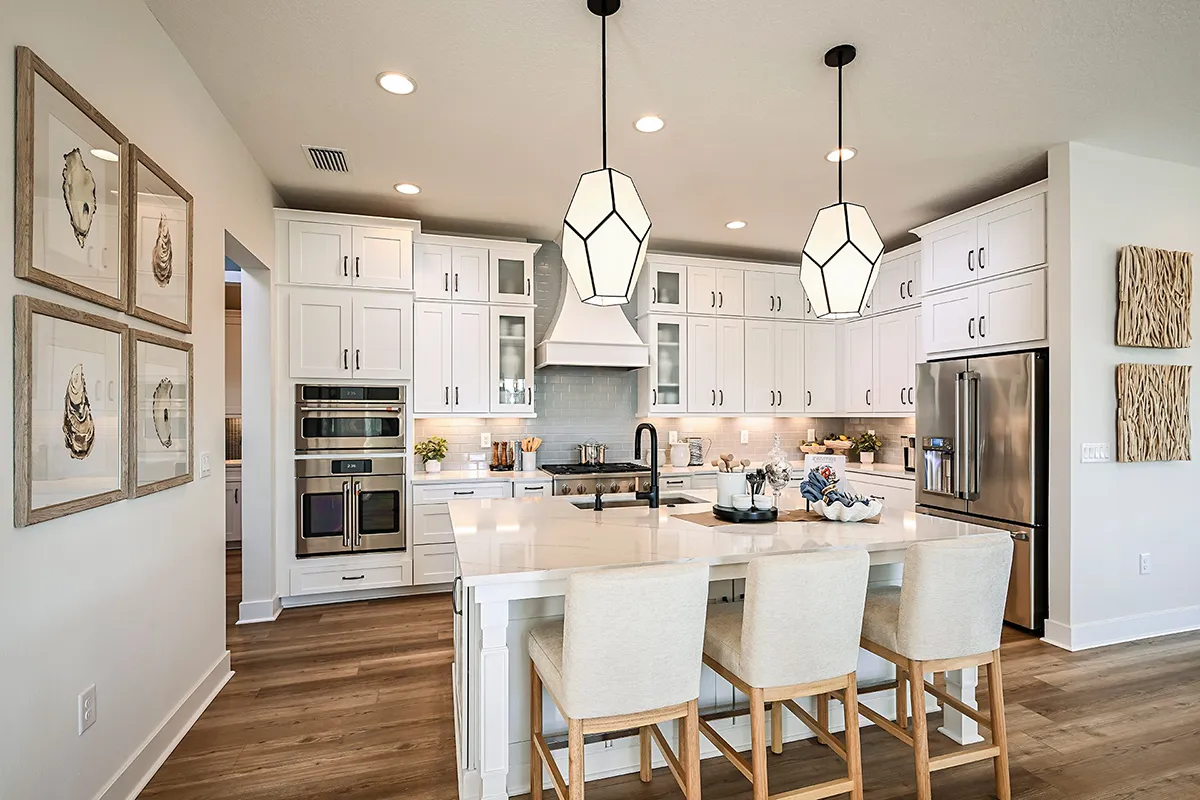 A modern kitchen with white cabinets, stainless steel appliances, a large island with three beige chairs, geometric pendant lights, and wood flooring. Art and decor hang on the light-colored walls.