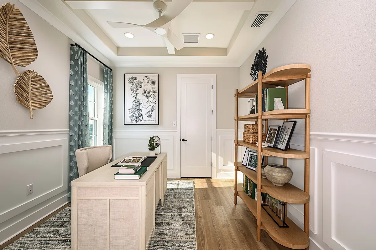 A modern home office with a desk, a wooden bookshelf holding decor and books, wall art, woven leaf wall decor, green curtains, and a ceiling fan. Natural light streams in through a window.