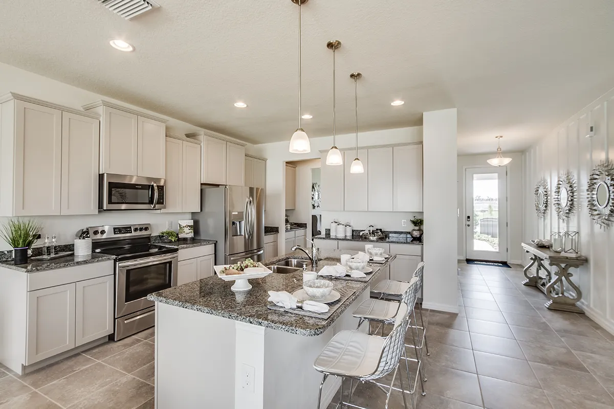 Modern kitchen with light gray cabinets, stainless steel appliances, granite countertops, a central island with stools, pendant lighting, and tile flooring. A hallway with a console table and mirrors is visible in the background.