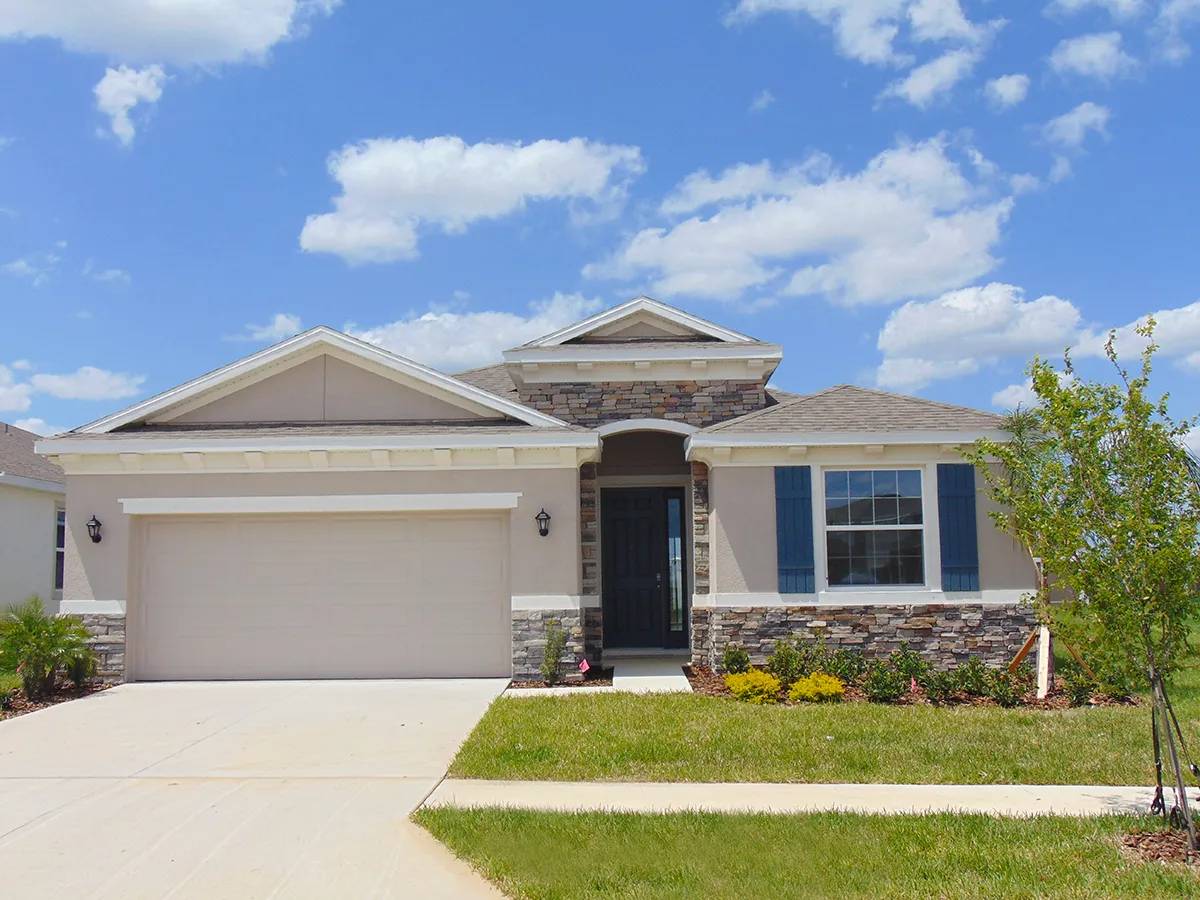 Single-story suburban house with a two-car garage, stone accents, blue shutters, and a black front door. The home has a well-kept lawn, small shrubs, and a partly cloudy sky in the background.