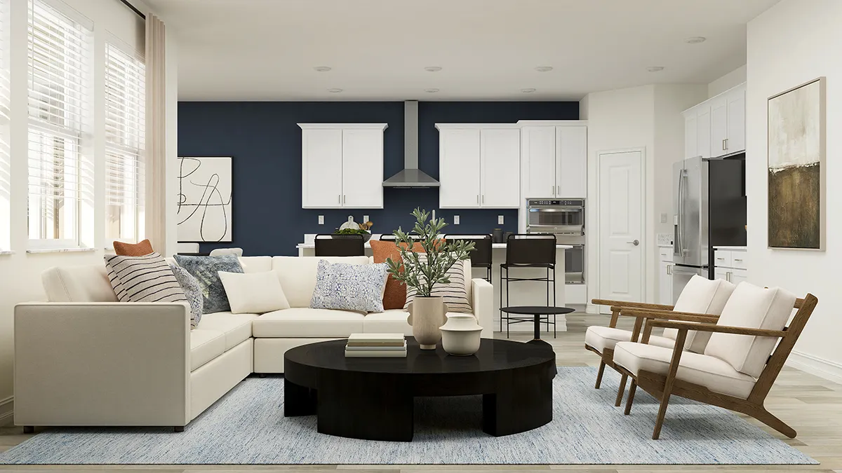 Modern living room with a white sectional sofa, wooden armchair, black round coffee table, and blue rug; kitchen with white cabinets and bar stools in the background. Large windows let in natural light.