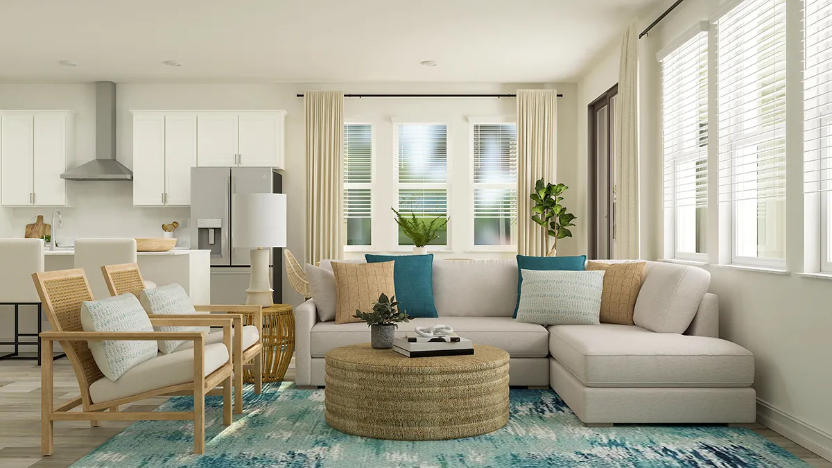 A modern living room with a beige sectional sofa, blue and neutral throw pillows, a round wicker coffee table, two wooden chairs, and a blue patterned rug. Large windows and a kitchen are visible in the background.