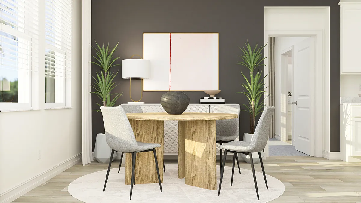 A modern dining area with a round wooden table, four gray chairs, a black vase centerpiece, two tall potted plants, a white sideboard, abstract wall art, and neutral walls with natural light from nearby windows.
