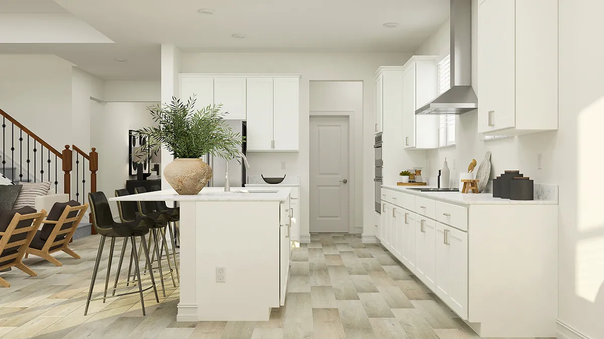 Modern kitchen with white cabinets, a central island with bar stools, stainless steel appliances, potted plant on the counter, and light wood flooring. Living area with chairs and staircase visible in the background.