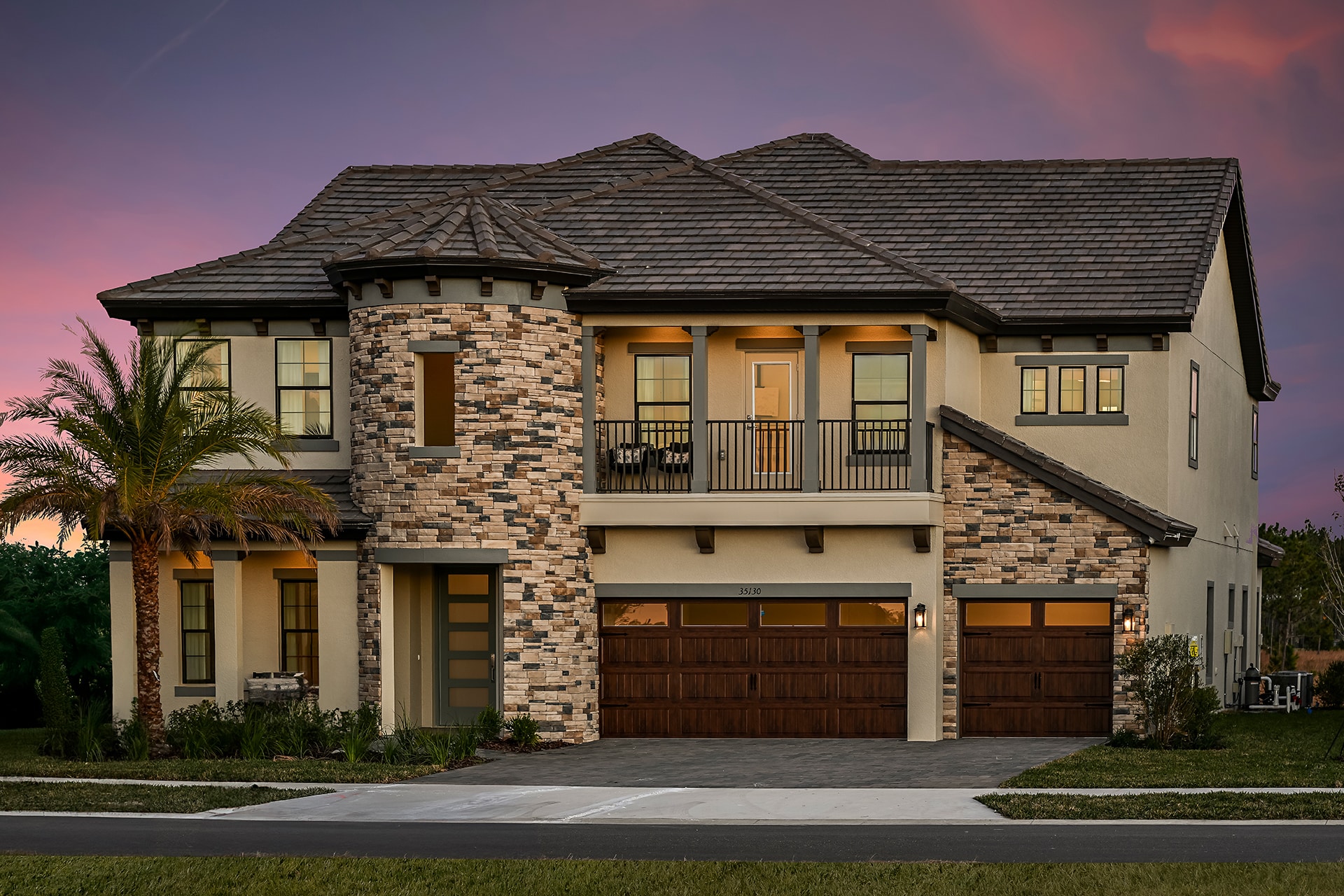 Two-story modern house with stone and stucco exterior, large windows, a wooden double garage door, a palm tree in front, and warm interior lights glowing at sunset.