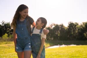 Two young girls hugging while standing in a sunlit grass field surrounded by trees.