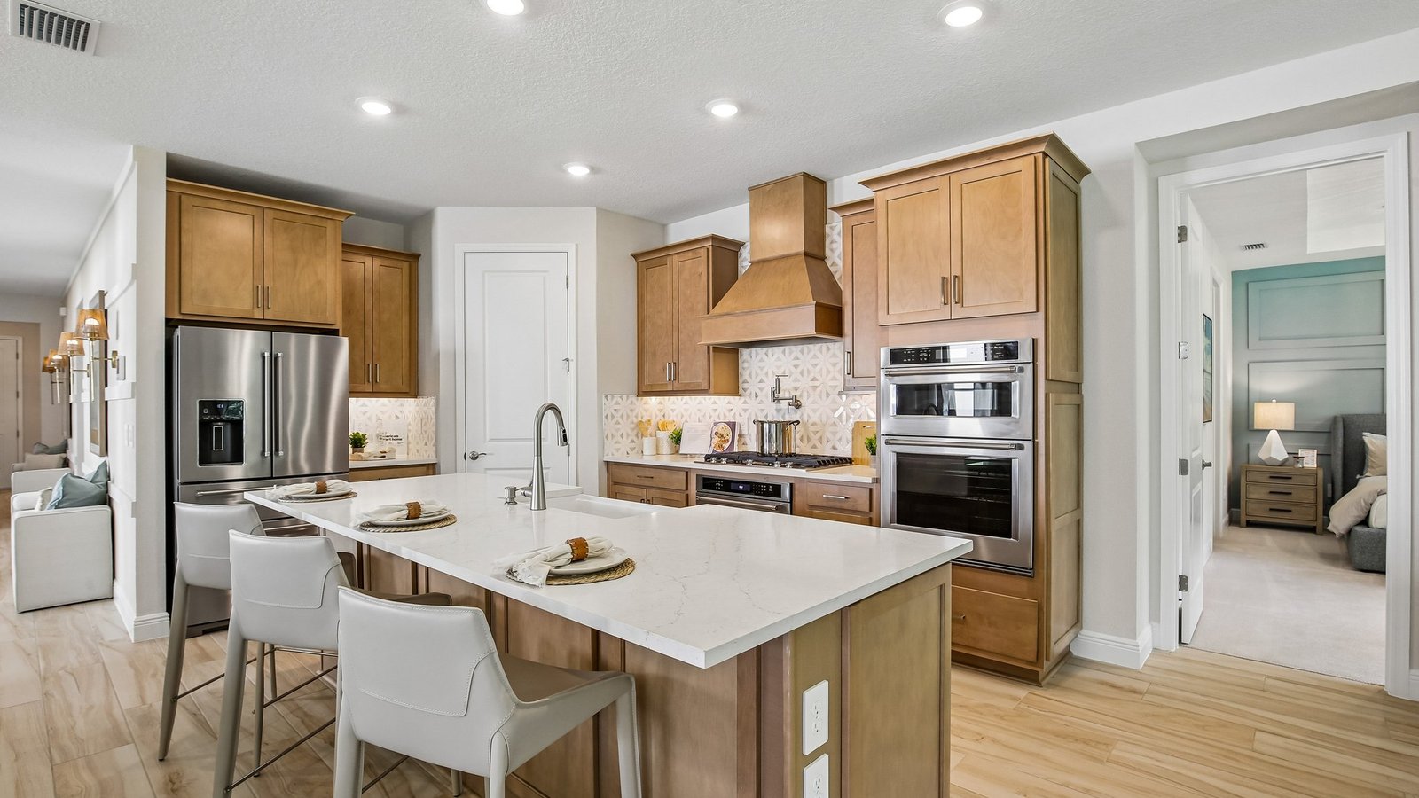 Modern kitchen with light wood cabinets, stainless steel appliances, white marble island with three place settings, beige barstools, and bright recessed lighting. Bedroom visible through doorway on the right.