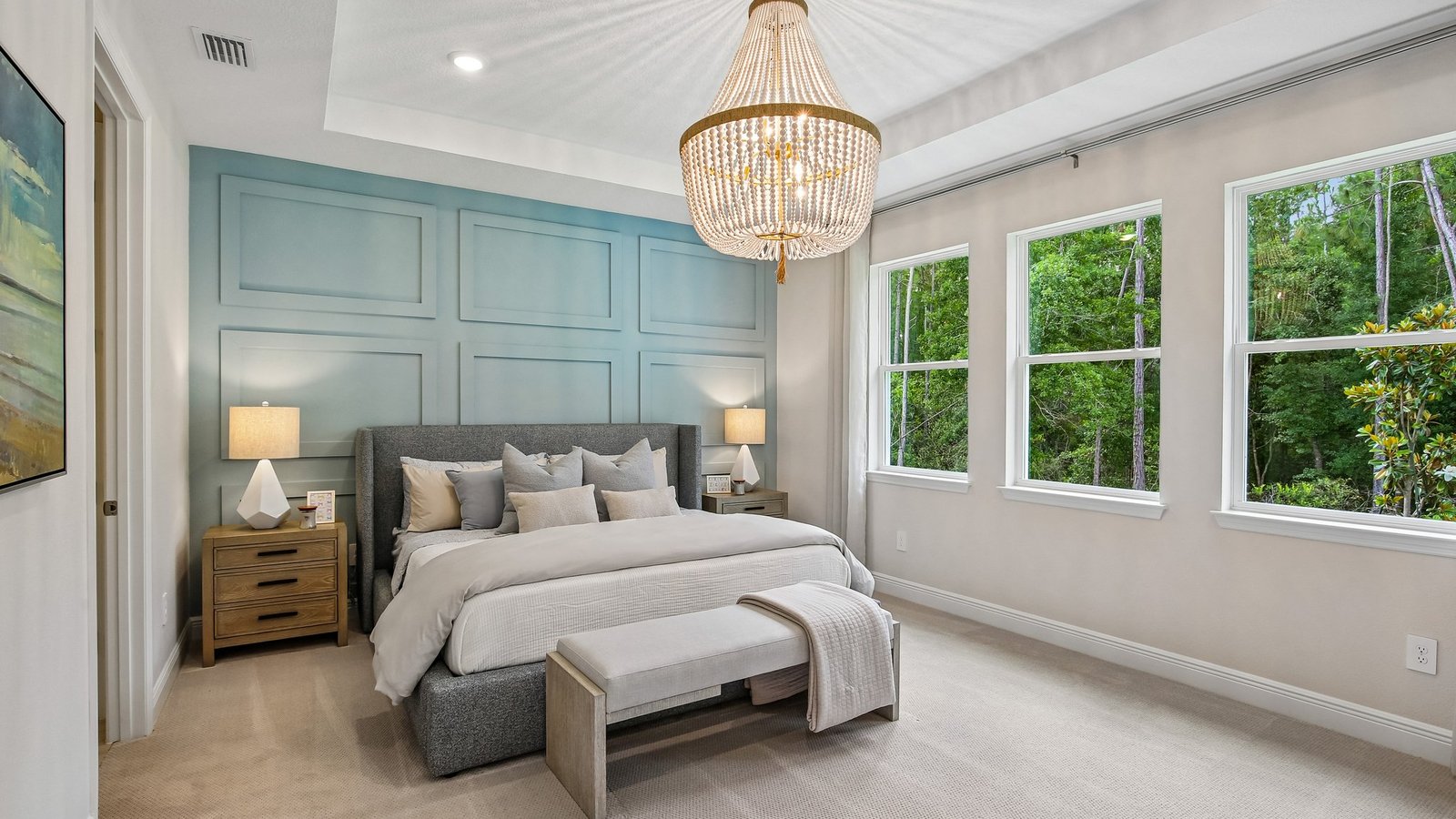 A modern bedroom with a large bed, gray upholstered headboard, beige bench, nightstands with lamps, a blue accent wall, three large windows, and a chandelier. Sunlight streams in, illuminating the neutral decor.