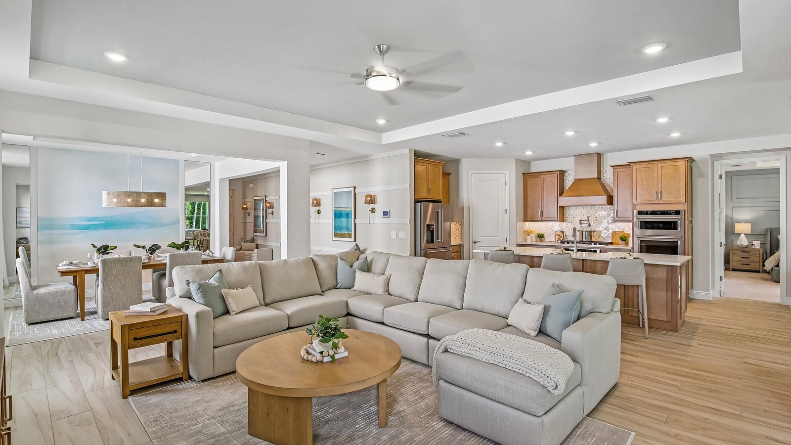Spacious open-concept living area with a large beige sectional sofa, round wooden coffee table, and modern kitchen with wood cabinets and stainless steel appliances; dining area visible in the background.