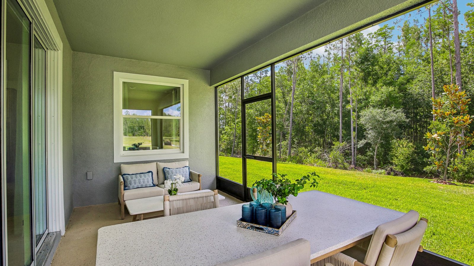 A screened-in patio with cushioned chairs, a white table with a decorative tray and plant, overlooking a green lawn and tall trees on a sunny day.