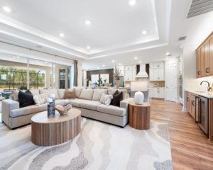 Modern open-concept living room and kitchen with a large beige sectional sofa, round wooden tables, neutral decor, wood flooring, and white cabinetry; large windows let in natural light from outside.