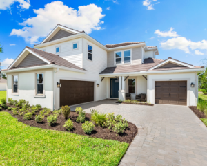 A modern two-story house with white exterior walls, brown garage doors, and a neatly landscaped front yard, set under a blue sky with scattered clouds. A paved driveway leads to two garages.