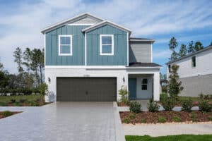 A modern two-story house with blue and white siding, a double garage, and a paved driveway. The front yard has green grass, shrubs, and small trees under a partly cloudy sky.