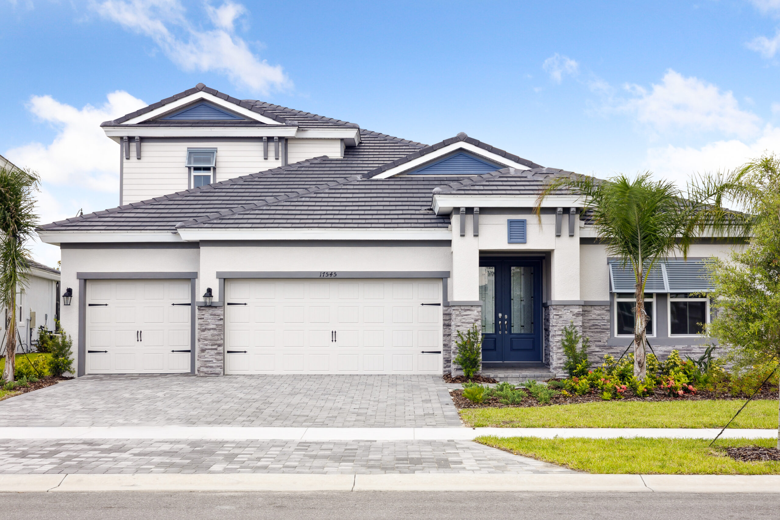 Modern two-story house with a gray tiled roof, white exterior, and stone accents. There are two garage doors, a dark blue front door, palm trees, and a paved driveway leading to the street.