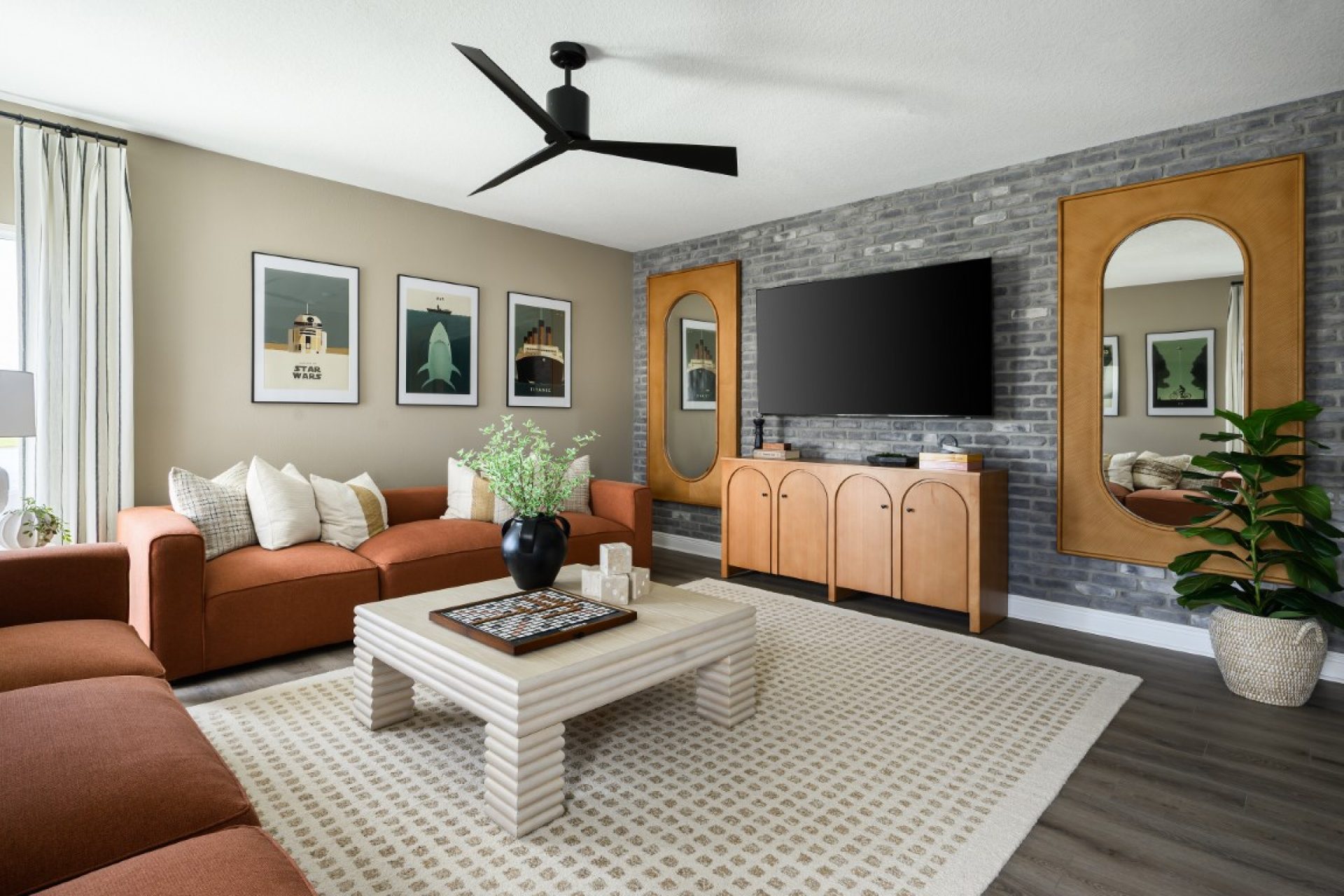 Modern living room with a rust-colored sofa, patterned rug, square coffee table, wall-mounted TV, two tall mirrors, framed art, and a potted plant against a gray brick accent wall.