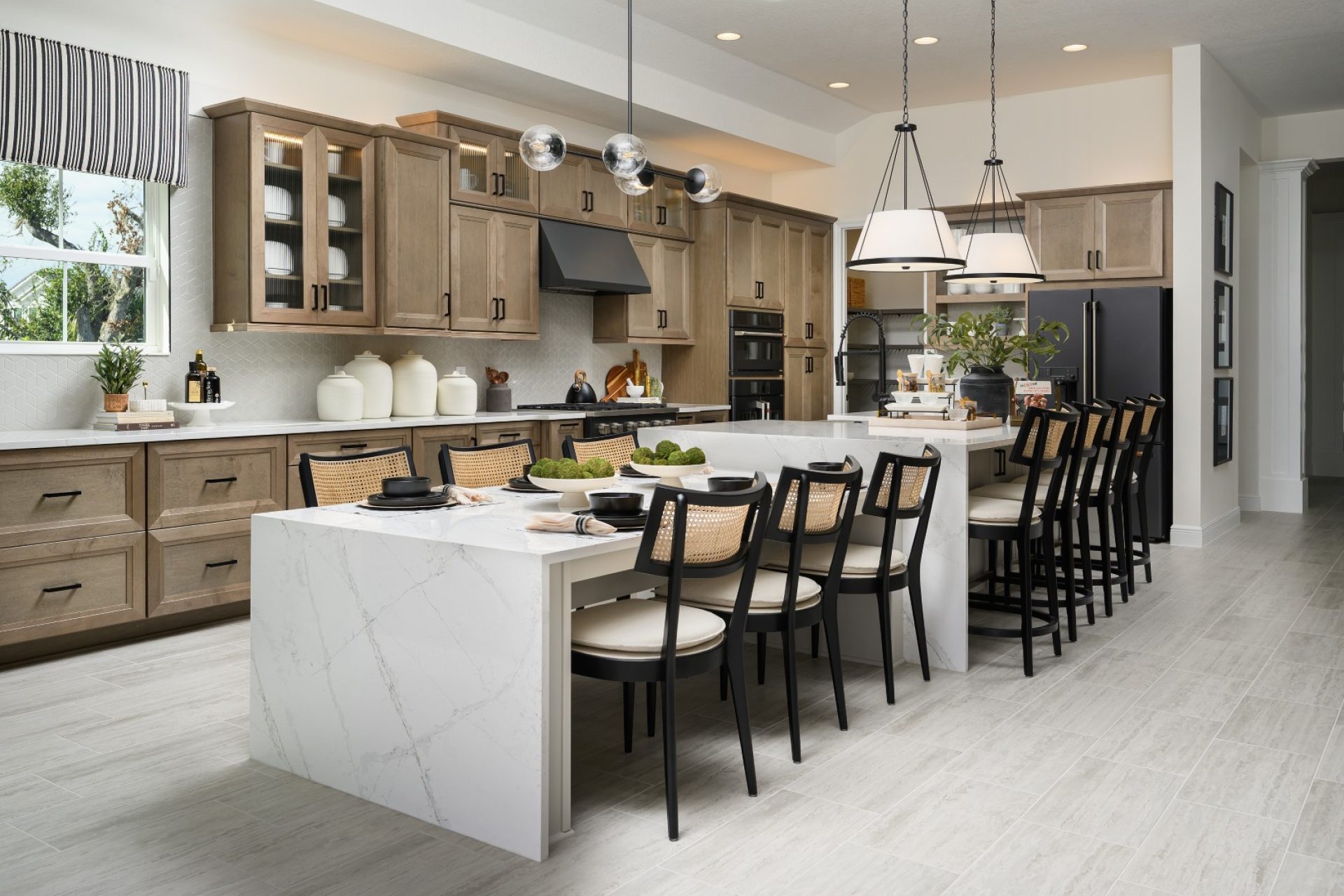 Modern kitchen with light wood cabinets, a large white marble island, black chairs, pendant lights, and stainless steel appliances. The island is set for dining, and greenery decorates the space. Light tile flooring throughout.