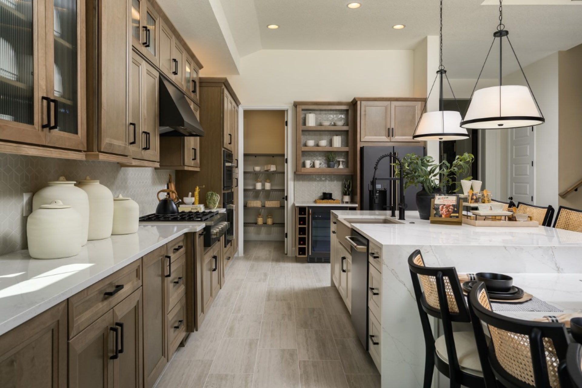 Modern kitchen with wood cabinets, white countertops, stainless steel appliances, a large island with black chairs, pendant lights, decorative jars, and open shelving displaying dishes and plants.