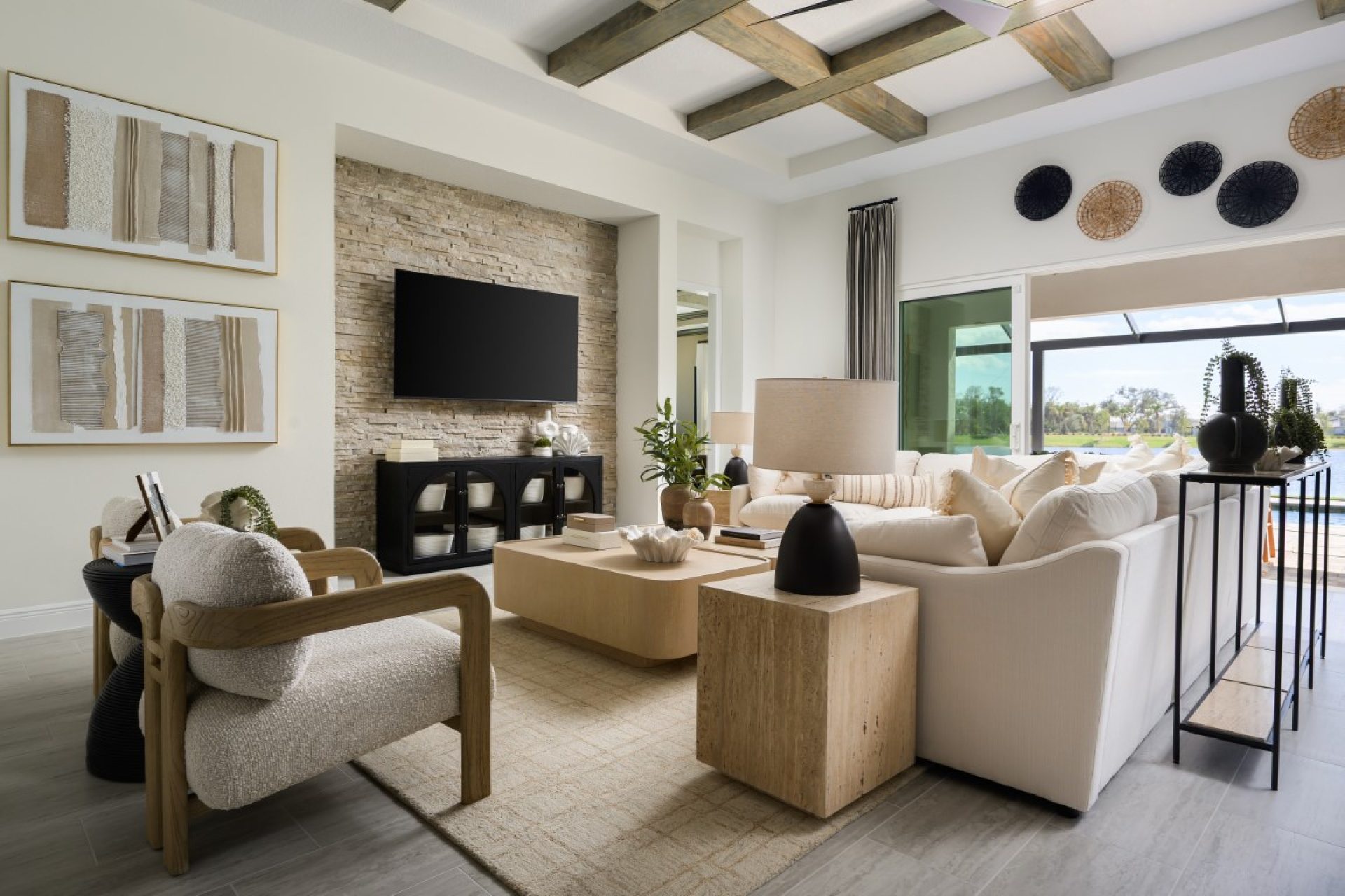 Modern living room with neutral tones, a white sectional sofa, wooden furniture, armchairs, a wall-mounted TV on a stone accent wall, large windows, and decorative wall art. Outdoor view visible through sliding glass doors.