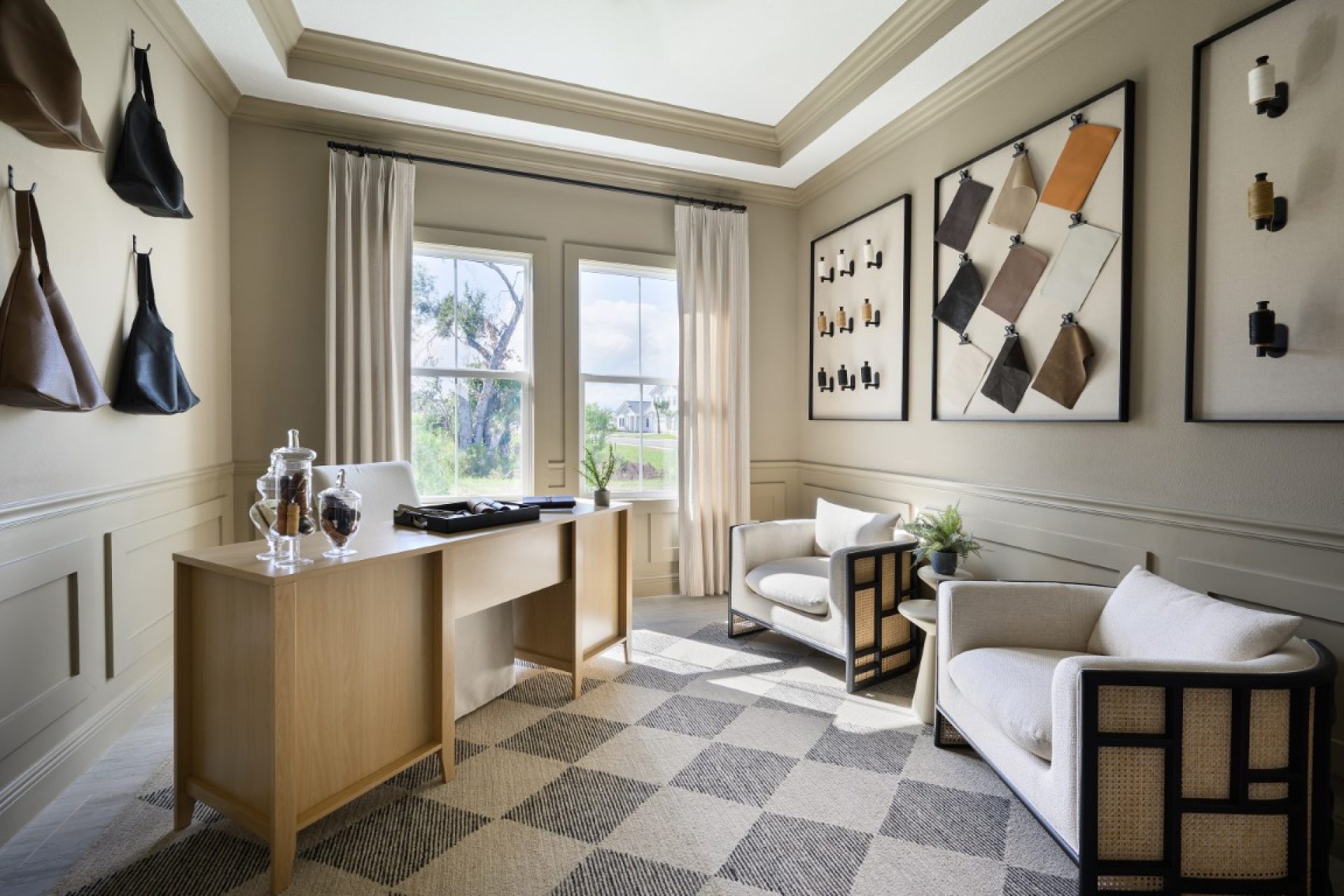 A stylish office with a light wood desk, two cream armchairs, large windows, patterned rug, and modern art on beige walls. Decorative bags and vases add a touch of elegance to the bright, inviting space.