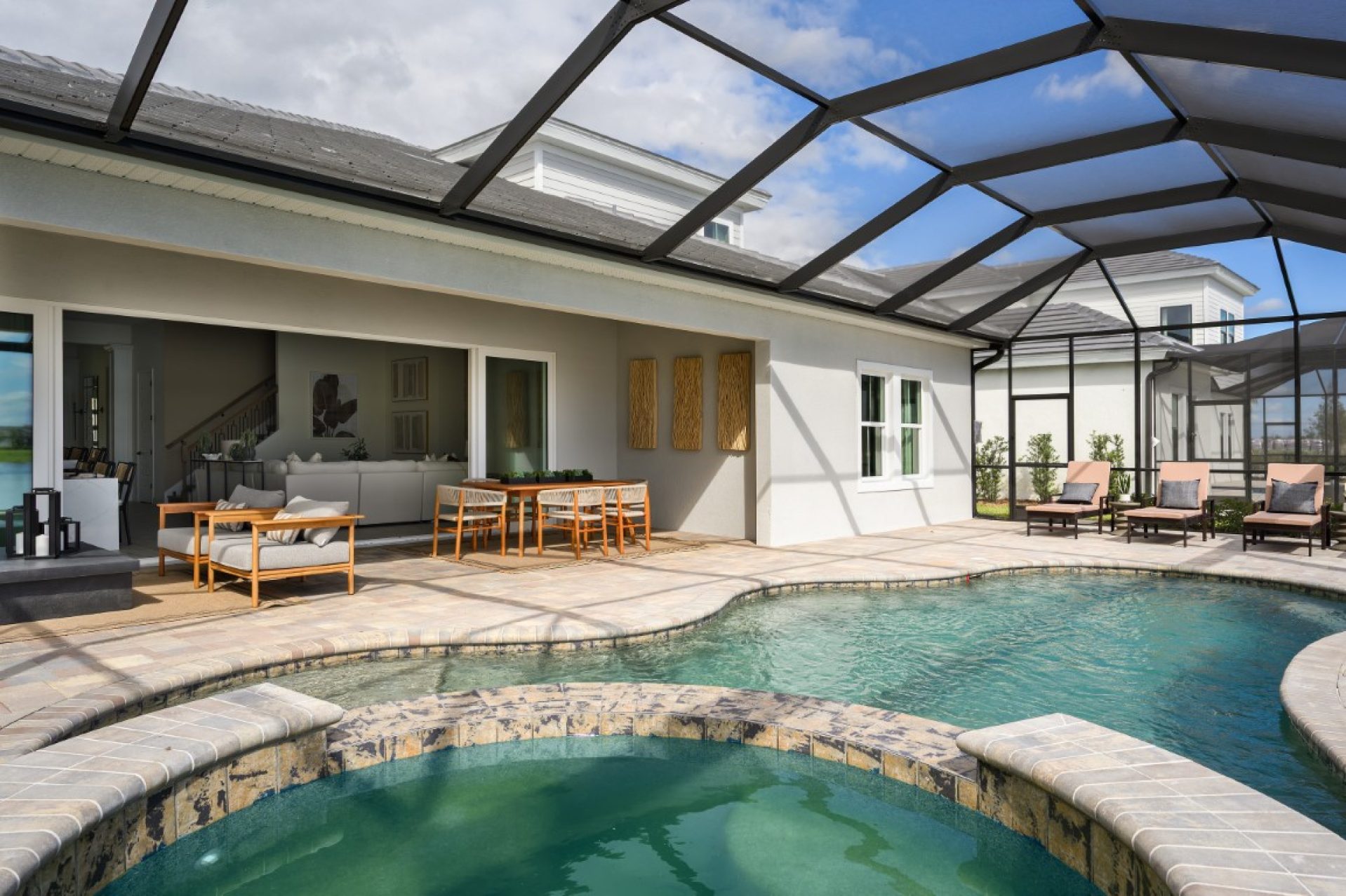 A modern screened-in patio with a swimming pool, hot tub, lounge chairs, a dining table, and an open living area visible through sliding glass doors under a partly cloudy sky.