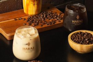 Three glasses of coffee drinks, including iced coffee and cold brew, sit on a dark table with scattered coffee beans. A wooden bowl and a cutting board hold more coffee beans. All glasses are labeled Haraz Coffee House.