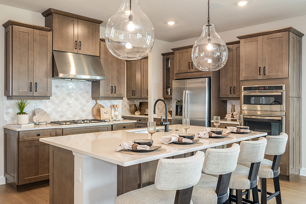 Modern kitchen with wood cabinets, stainless steel appliances, double ovens, and a large island with four cushioned chairs. Pendant lights hang above the island, which is set with plates, napkins, and glasses.